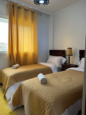 A hotel room with two beds, a window with yellow curtains, a bedside lamp, and a view of the outside. - SINIM HOUSE (Taghazout)