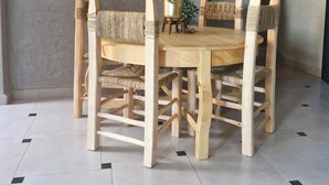 A dining area with a round wooden table and four chairs, each with woven seats. A round mirror with a wooden frame hangs on the wall above the table. - SINIM HOUSE (Taghazout)