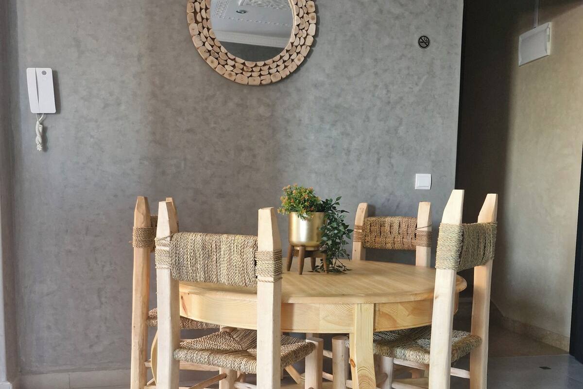 A dining area with a round wooden table and four chairs, each with woven seats. A round mirror with a wooden frame hangs on the wall above the table.