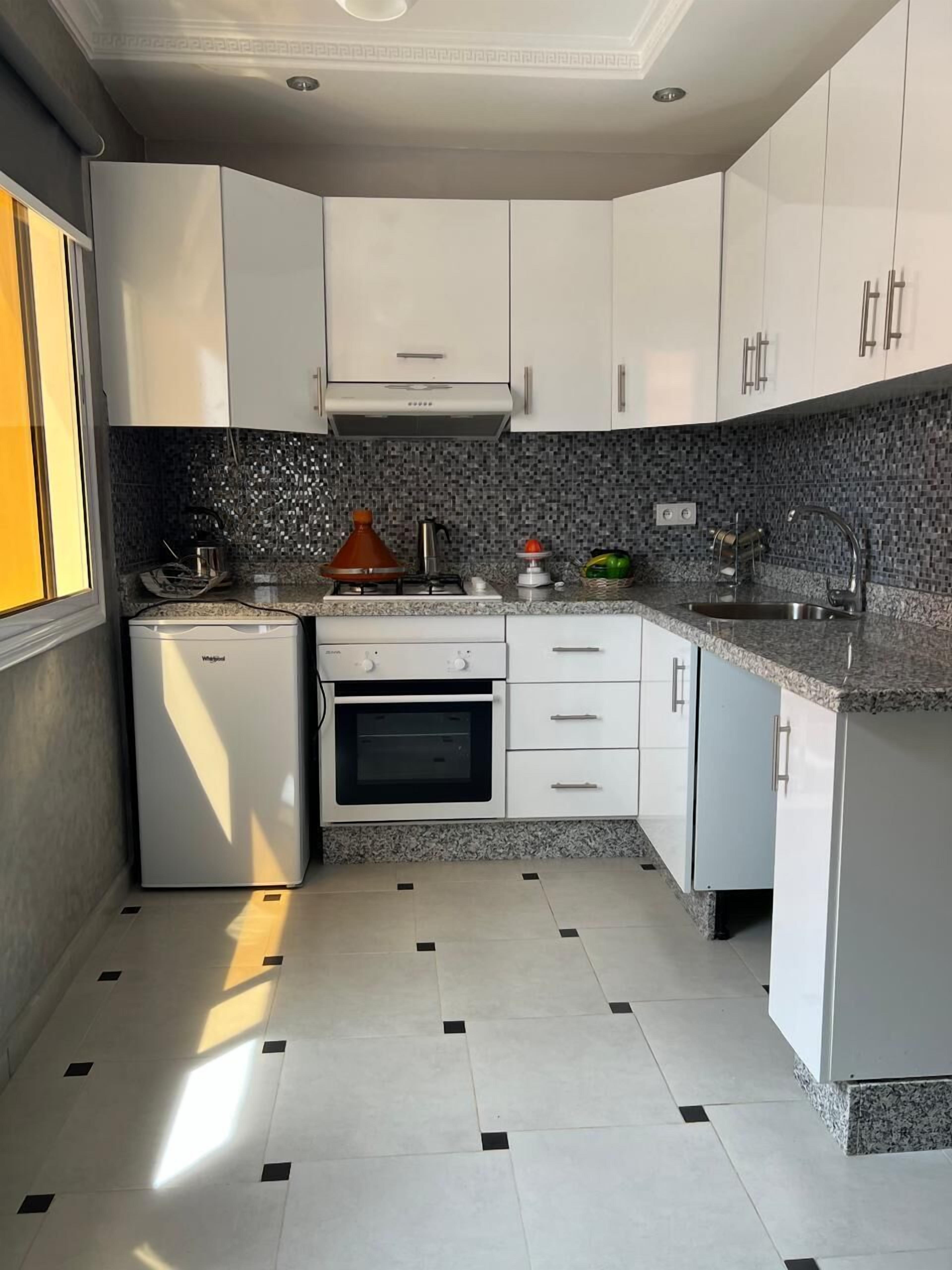 Cozinha compacta com armários brancos, forno embutido, geladeira pequena e painel de azulejos.