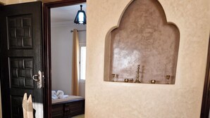 A room with a wooden chair, a white box with a decorative design, a dark door, and a wall-mounted shelf with small items. - SINIM HOUSE (Taghazout)