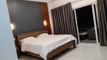 Habitación de hotel con cama de madera, mesita de noche y puerta corredera de cristal que da a un área exterior con mobiliario de mimbre.