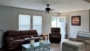 A spacious living room with a brown leather sofa, a brown armchair, and a beige ottoman. There is a glass coffee table with decorative vases. The room has wooden flooring and a ceiling fan. - Your Toledo Bend Lakefront Home – Neighborhood Boat Launch Just Next Door (Many)