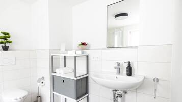 A modern bathroom with a white sink, toilet, and a mirror above the sink. There is a small plant on the sink and a toilet paper holder on the wall.