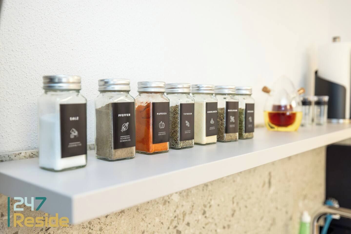 Ein Regal mit verschiedenen Gewürzgläsern, beschriftet mit Bezeichnungen wie 'Pfeffer' und 'Paprika'.