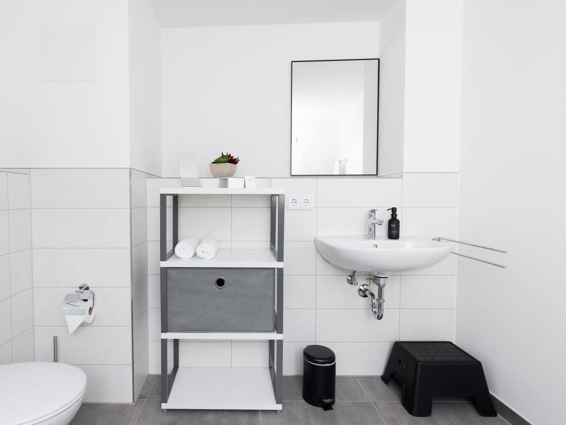 Une salle de bain moderne avec des toilettes blanches, un meuble de rangement gris, un lavabo blanc et une poubelle noire.