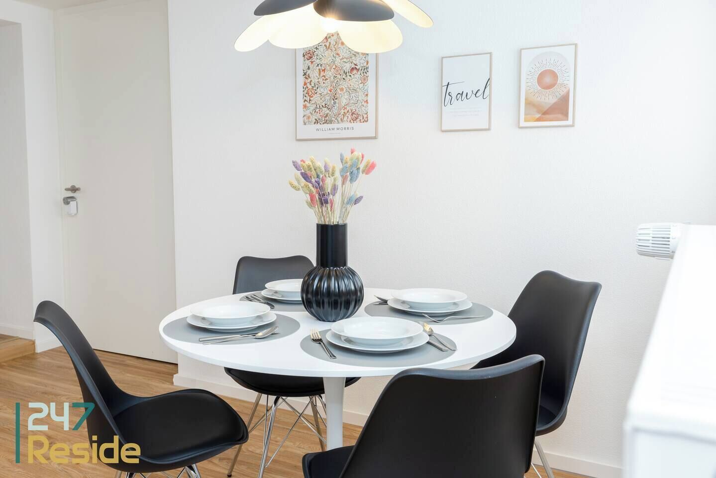 A dining area with a round table, black chairs, a black vase with flowers, and framed artwork on the wall.