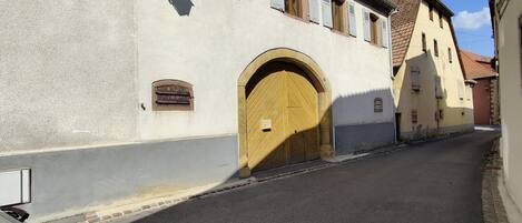 Una calle estrecha con un edificio que tiene una puerta de madera grande y arqueada, y una ventana pequeña con persianas.