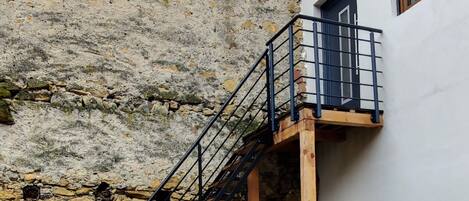 A stone building with a wooden staircase leading to a balcony. There is a small table and two chairs outside.