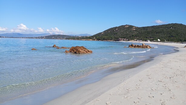 Ein Strand mit klarem, blauem Wasser, sandigem Ufer und Felsformationen.