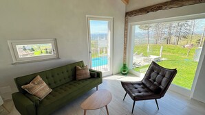 A spacious living room with a green sofa, a brown leather chair, and a wooden coffee table. There is a large window offering a view of a pool and a green landscape. - Spectacular views of UNESCO Castles, Langhe valley, French, Italian & Swiss alps (Costigliole di Asti)