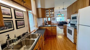 Una cocina con armarios de madera, un lavamanos de acero inoxidable, un microondas y un horno. Hay cuadros en la pared y una ventana con persianas.