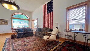 Una sala de estar con una gran bandera estadounidense, dos sofás estampados, un sillón blanco, una mesa de comedor con sillas y un tapete grande.