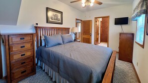 Un dormitorio con cama, cómoda de madera, ventilador de techo, cuadro enmarcado y televisor en la pared.
