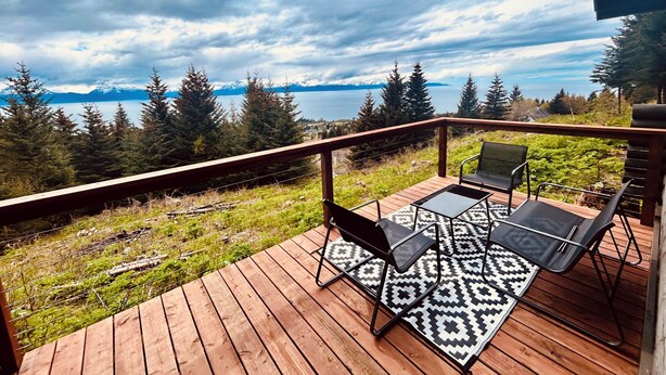 A wooden deck with outdoor furniture and a patterned rug, overlooking a lake and mountains. - Cozy home with a view, newly remodeled. (Homer)