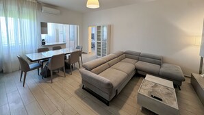 A modern living room with a sectional sofa, a dining table with chairs, and a large window with sheer curtains. - Suite Patrizia - Appartamento nel Cuore di Modena (Modena)