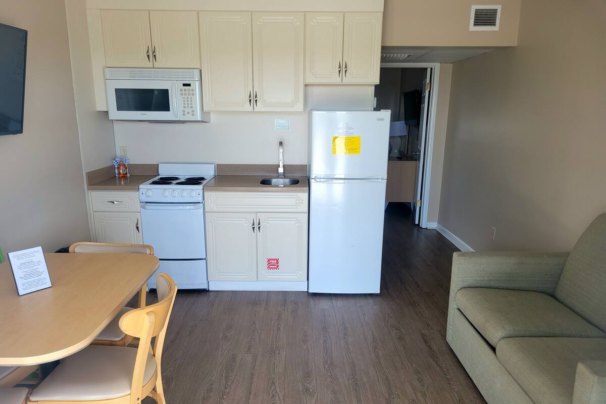 A compact kitchen with a microwave, stove, sink, and refrigerator. There is a dining table with chairs and a sofa in the living area.