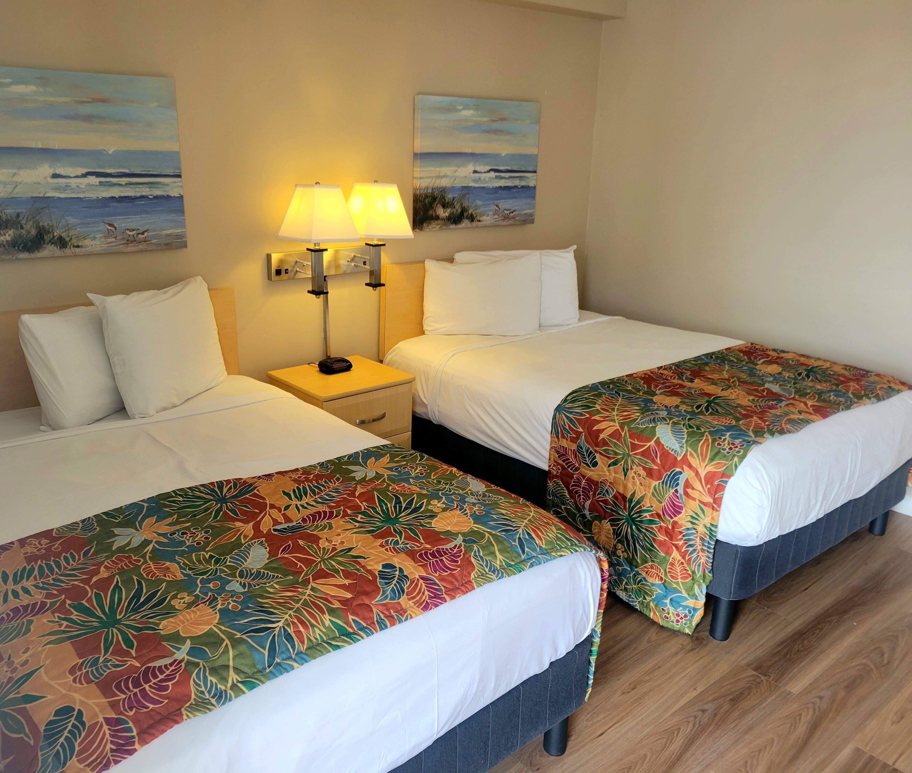 A hotel room with two beds, each with a floral bedspread, a nightstand with a lamp, and two wall-mounted paintings of a beach scene.