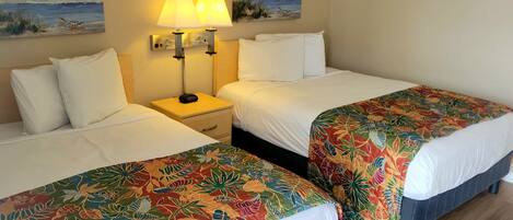A hotel room with two beds, each with a floral bedspread, a nightstand with a lamp, and two wall-mounted paintings of a beach scene.