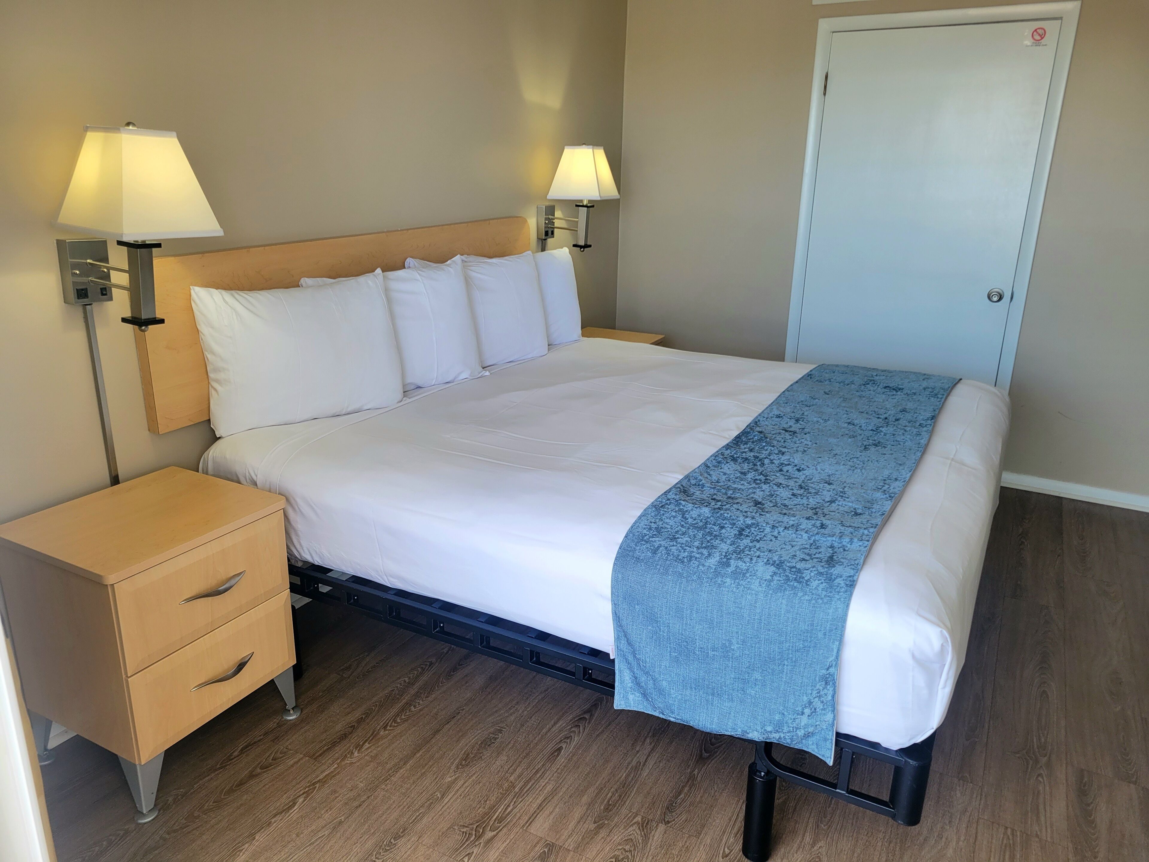 A neatly made bed with white linens and a blue blanket, flanked by two bedside tables with lamps, in a room with wooden flooring and a door in the background.
