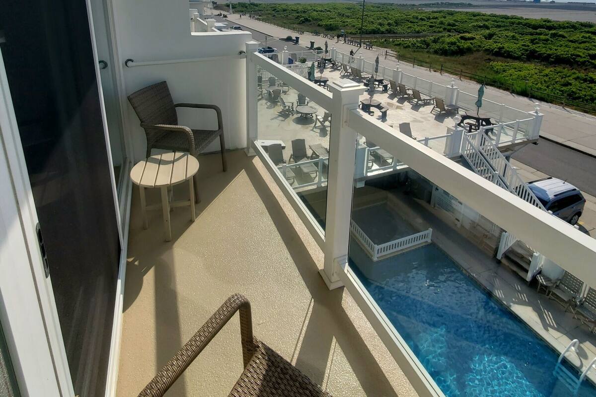 A balcony with a pool, wicker chairs, and a view of the beach and ocean.