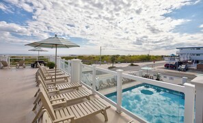 A poolside area with lounge chairs and umbrellas, a clear view of the ocean, and a building in the background. - Water's Edge Ocean Resort (Wildwood Crest)