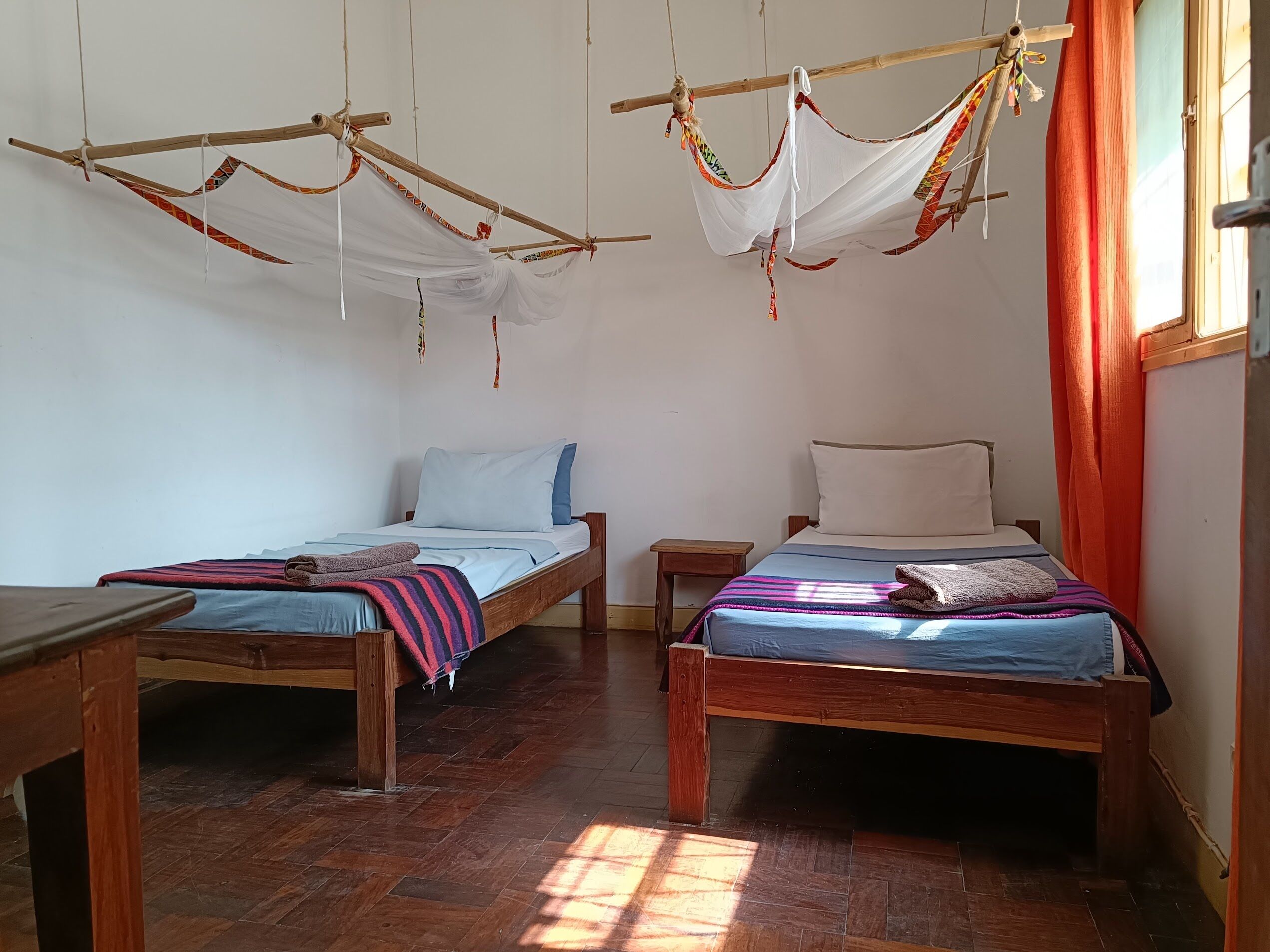 Quarto com duas camas de madeira, cada uma com travesseiro e cobertor. Há mesas de madeira e um pequeno banquinho. O chão é de azulejos e a janela tem cortinas laranjas.