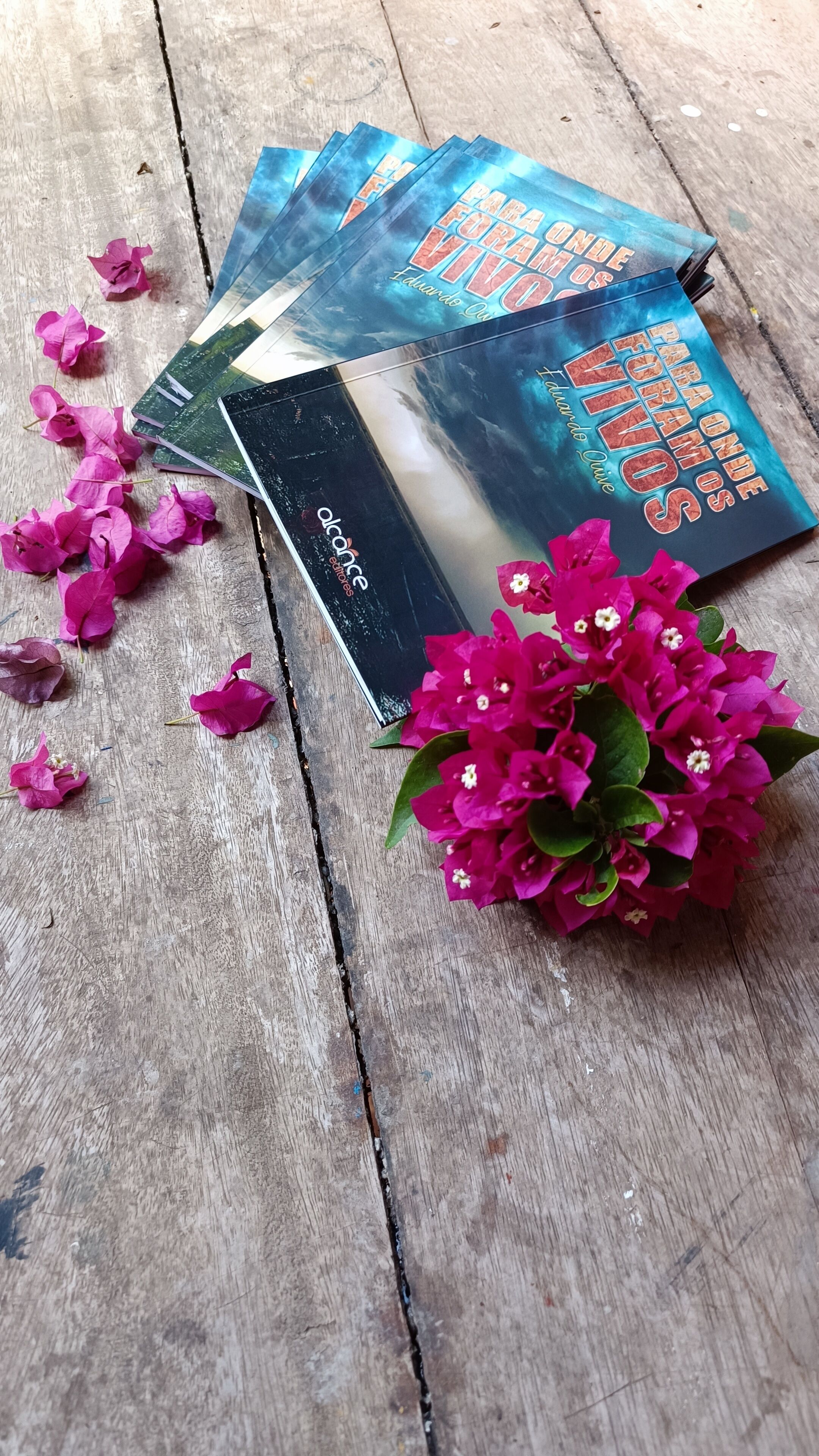 A wooden table with a stack of books titled "Para Onde Vamos" and a bouquet of pink flowers.