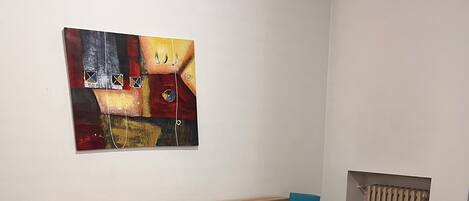 Habitación de hotel con cama de madera, dos mesitas de noche con lámparas, un radiador y un cuadro abstracto de colores en la pared.