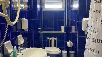 Une salle de bain avec un lavabo, des toilettes et un miroir. Il y a une fenêtre avec des stores, un porte-serviettes et un rideau de douche avec un motif imprimé.