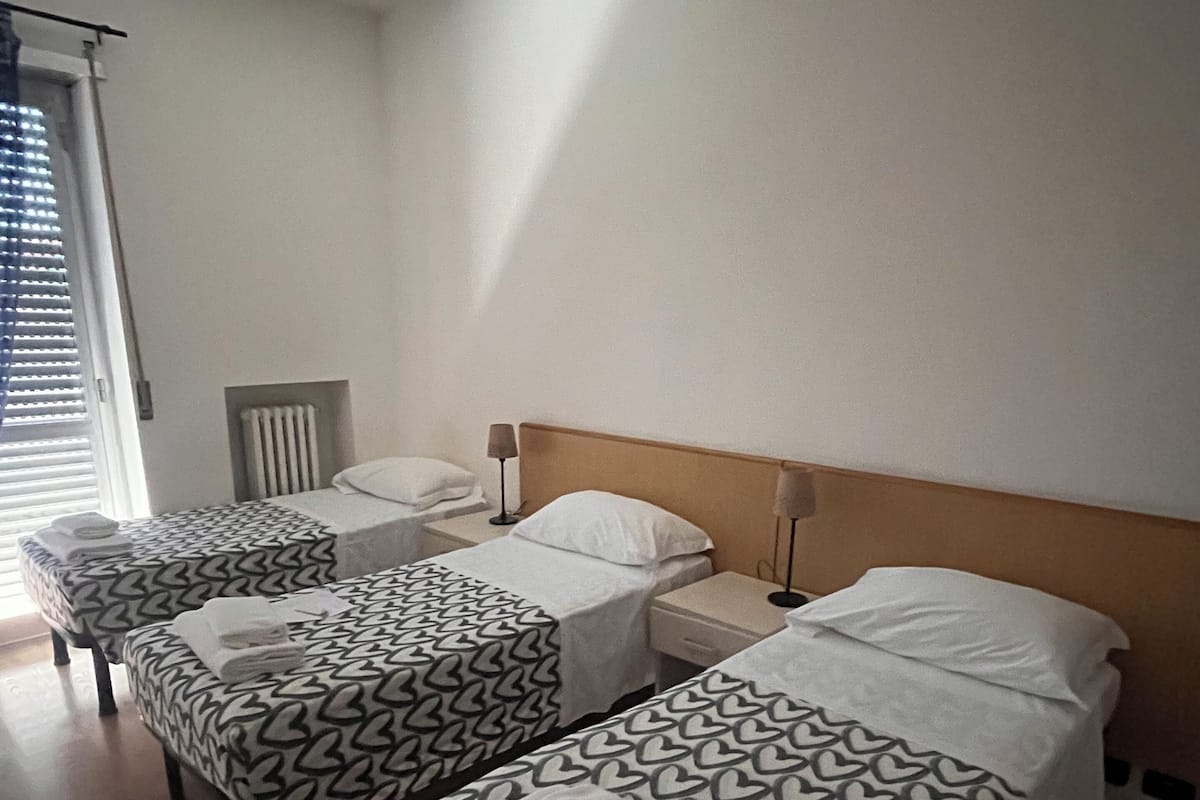 A hotel room with three single beds, each with a patterned bedspread and white pillows. There is a bedside table with a lamp on each bed, and a radiator on the wall.