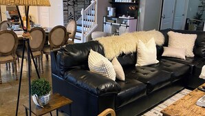Uma sala de estar moderna com um sofá de couro preto, almofadas brancas, uma mesa de centro de madeira e um espaço de refeições com cadeiras e um abajur.