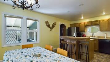 A dining area with a table covered in a floral tablecloth, a chandelier, and a kitchen with wooden cabinets and a refrigerator.