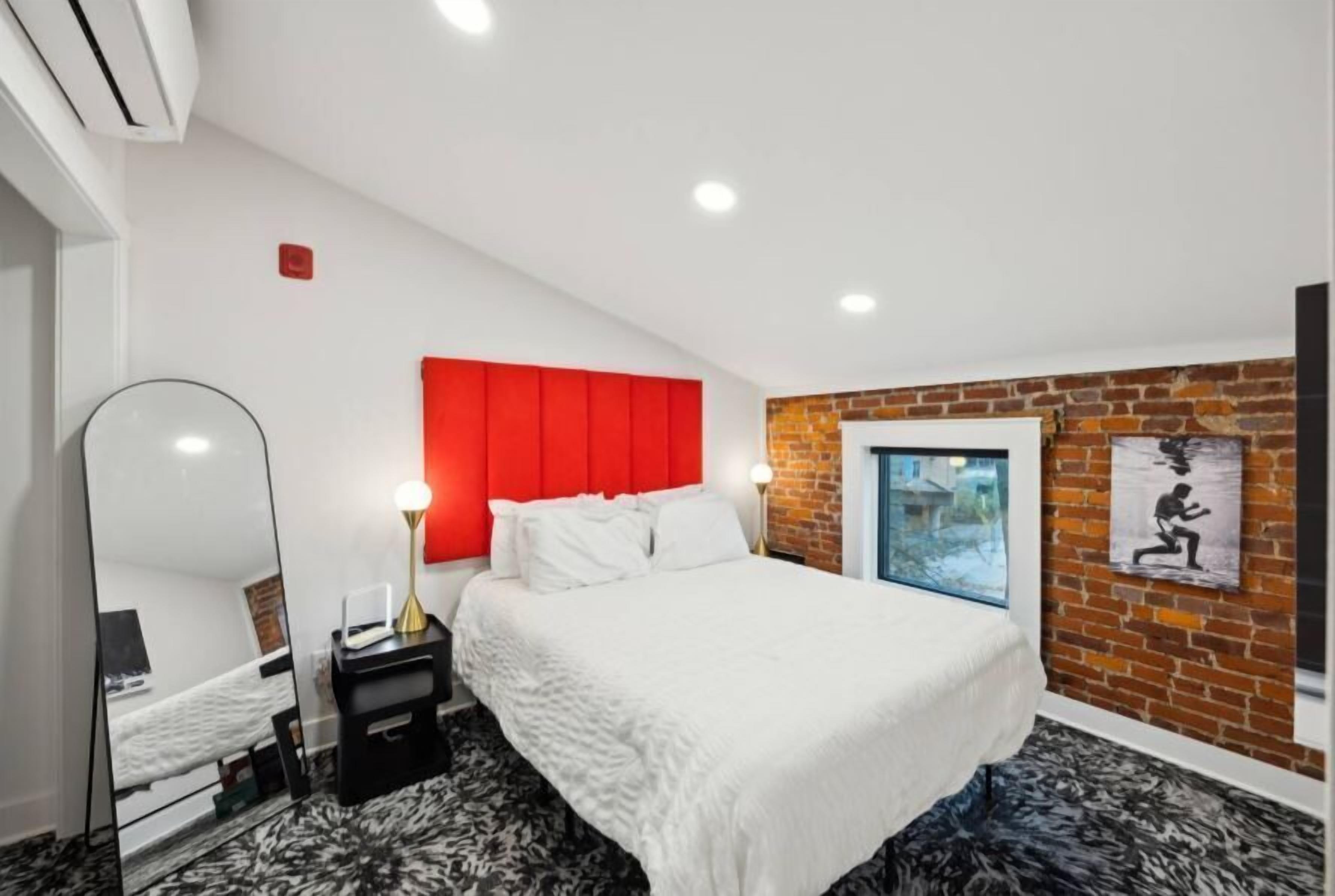 A modern bedroom with a red headboard, a large bed, a mirror, a nightstand, and a window with a view of houses.