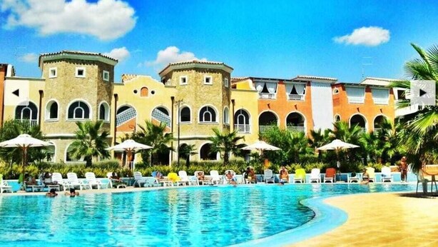 Un área de piscina con sillones de descanso y sombrillas, rodeada de edificios con ventanas de arco y balcones.