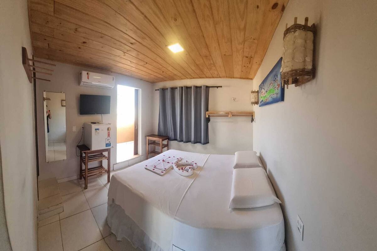 Un dormitorio con techo de madera, cama con ropa de cama blanca, televisor montado en la pared y ventana con cortinas.