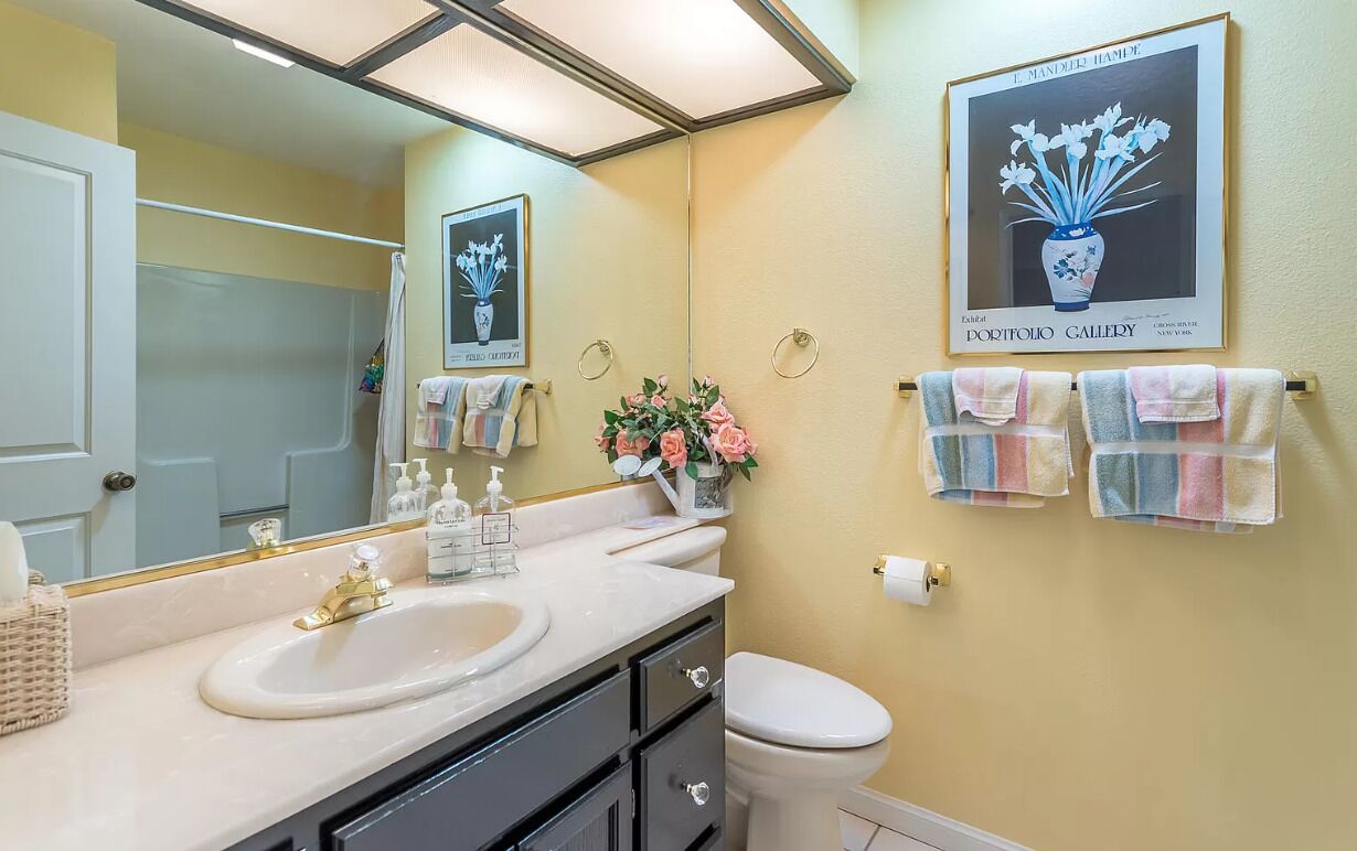 A bathroom with a large mirror, a sink, a toilet, and a shower. There are towels hanging on the wall and a framed picture above the sink.