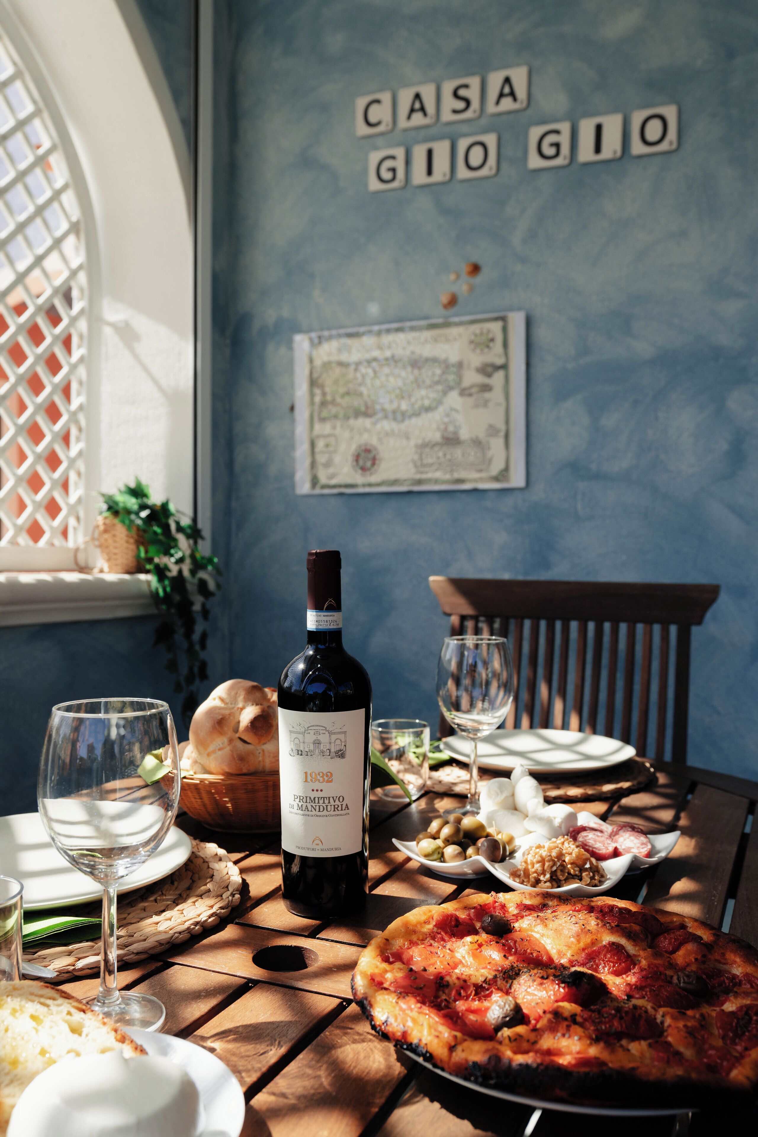 Un tavolo da pranzo con pizza, una bottiglia di vino e diversi piatti, su uno sfondo con una parete blu, una finestra e una mappa.