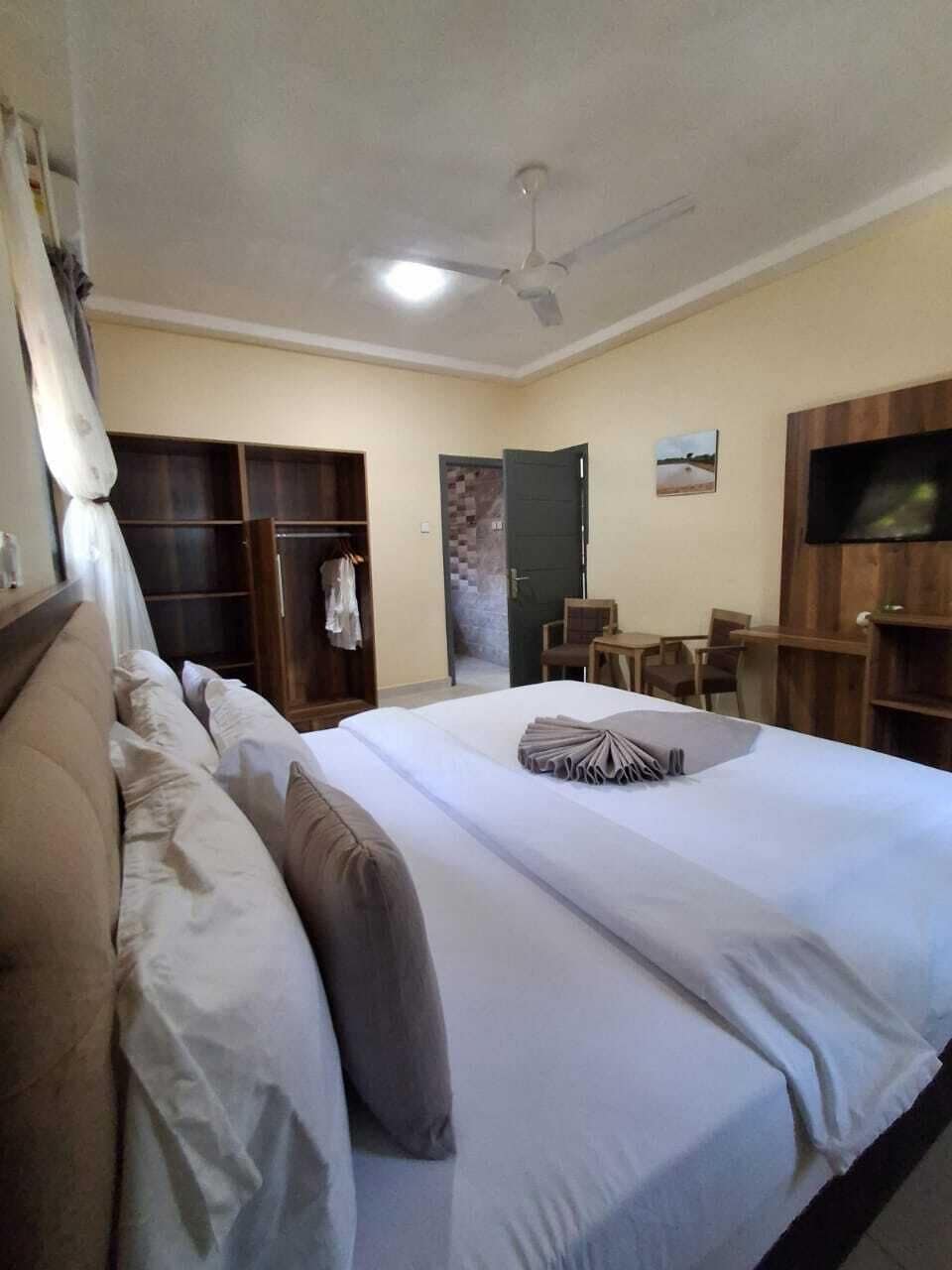 A hotel room with a large bed, a wardrobe, a desk with chairs, a television, and a ceiling fan.