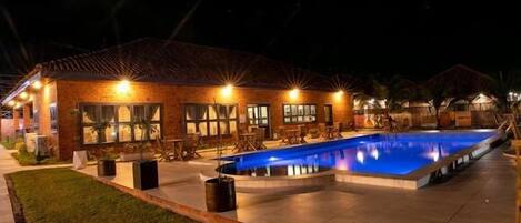 A nighttime view of a hotel with a swimming pool, outdoor seating, and a brick facade.