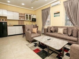 A living room with a sofa, armchair, and coffee table. There is a kitchen area with cabinets and a refrigerator.