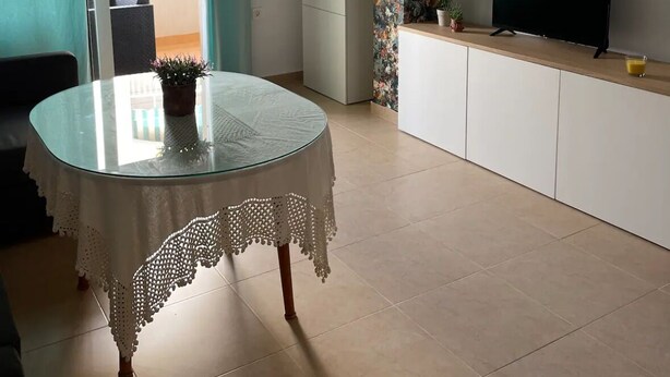 A modern living room with a glass table, a flat-screen TV, and a kitchen area with white cabinets.