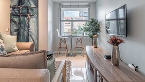 A modern living room with a large abstract painting, a sofa, a coffee table, a wooden side table, a potted plant, and a TV mounted on the wall. - KMG Apartments at The Apex on Smuts  (Johannesburg)