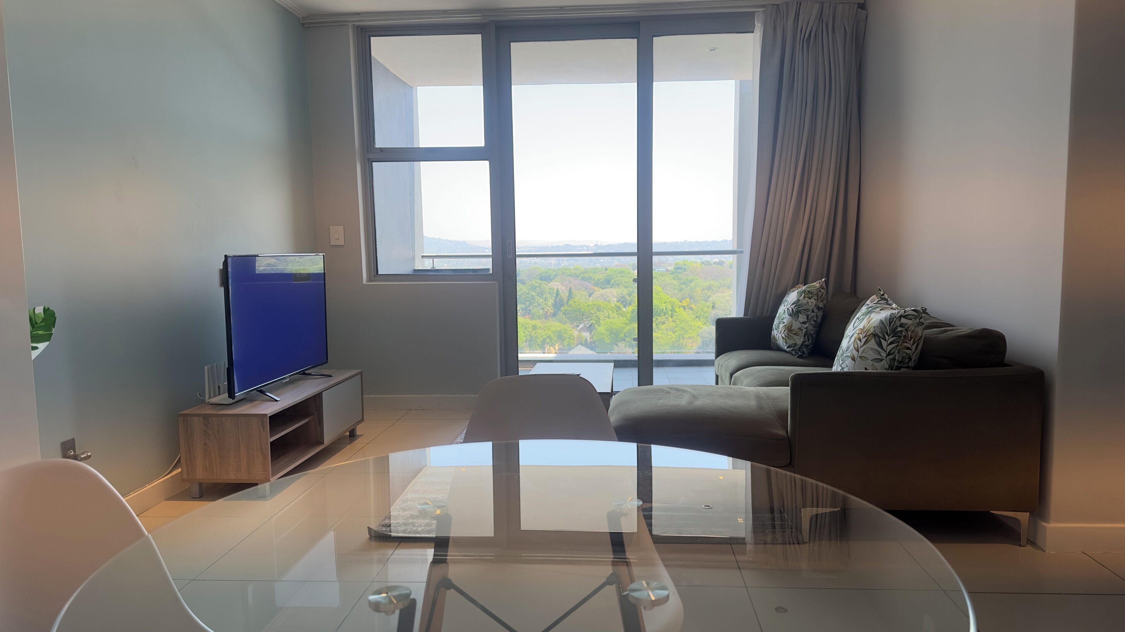 A modern living room with a glass coffee table, a flat-screen TV on a wooden stand, a grey sofa with patterned cushions, and a large window offering a view of greenery and buildings.