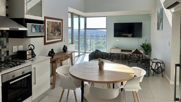 A modern kitchen with a dining area, large windows, and a flat-screen TV. - KMG Apartments at The Apex on Smuts  (Johannesburg)