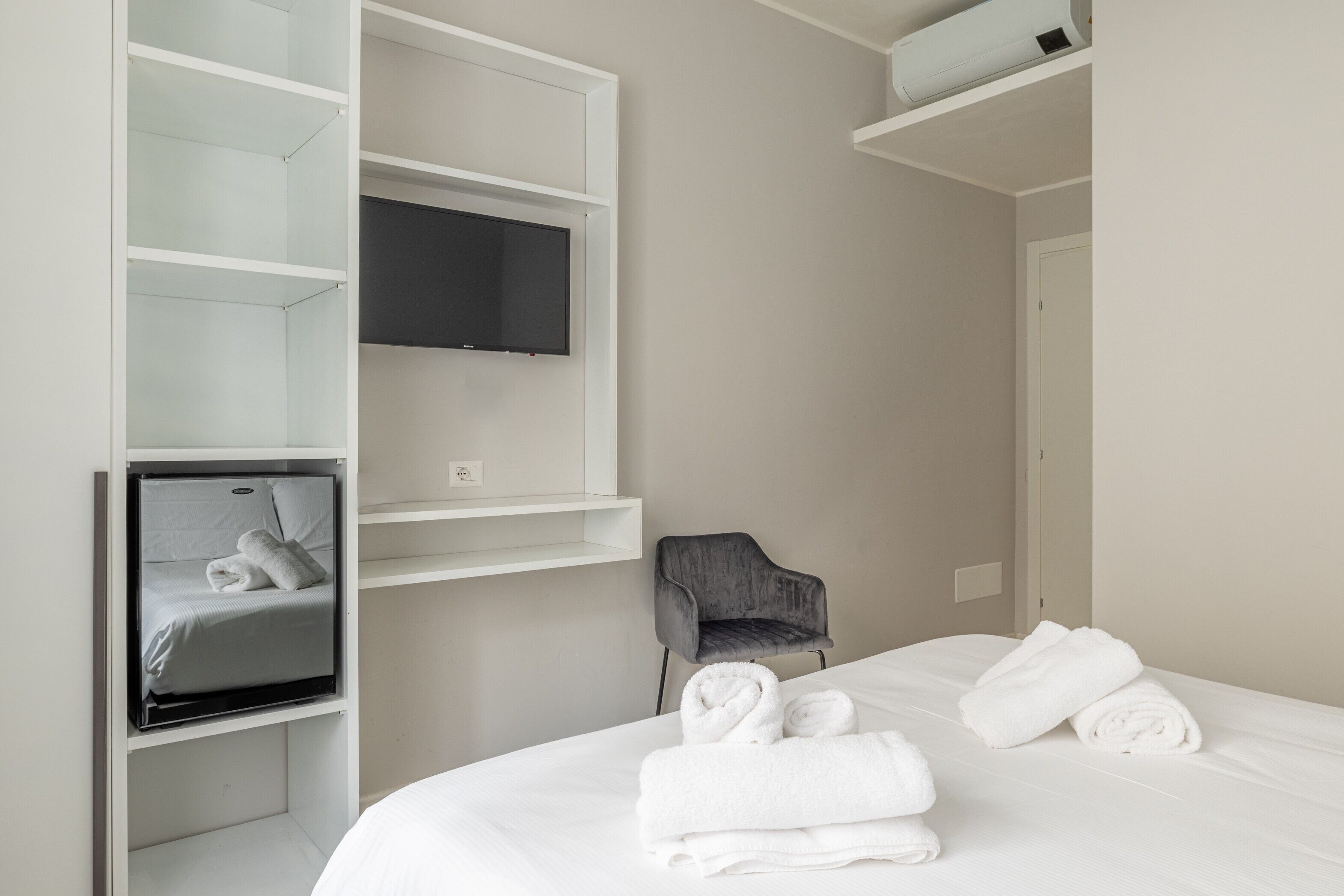A modern hotel room with a bed, a flat-screen TV mounted on a wall, a grey armchair, and neatly folded towels.