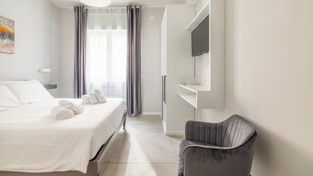 A modern hotel room with a neatly made bed, a grey upholstered chair, a flat-screen TV mounted on the wall, and a large window with curtains. - Belli Suites Vatican Rome Guest House (Rome)