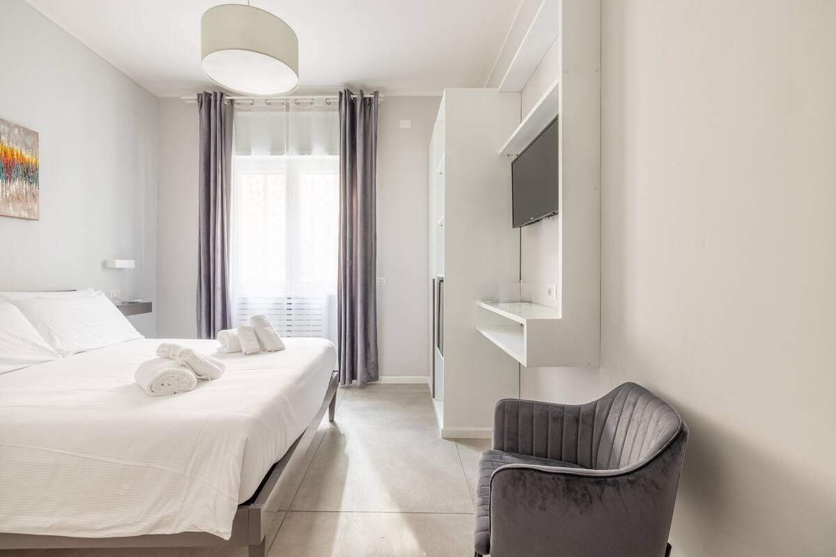 A modern hotel room with a neatly made bed, a grey upholstered chair, a flat-screen TV mounted on the wall, and a large window with curtains.
