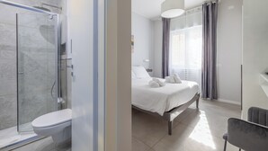 A modern bathroom with a glass shower enclosure, a toilet, and a bed with white linens. - Belli Suites Vatican Rome Guest House (Rome)