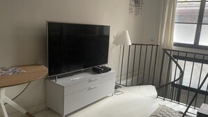 A modern living room with a flat-screen TV on a white cabinet, a small table with a magazine, a white lamp, and a view of the street through a window. - Duplexe Paris 6eme (Paris)